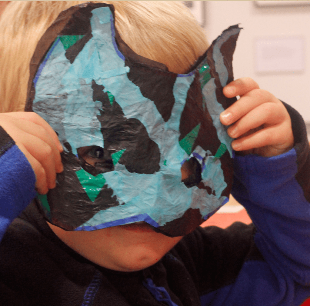 A child holding up a colourful, reclaimed plastic bag mask, with blue, black, and green shapes, covering their face while peeking through one of the eye holes.