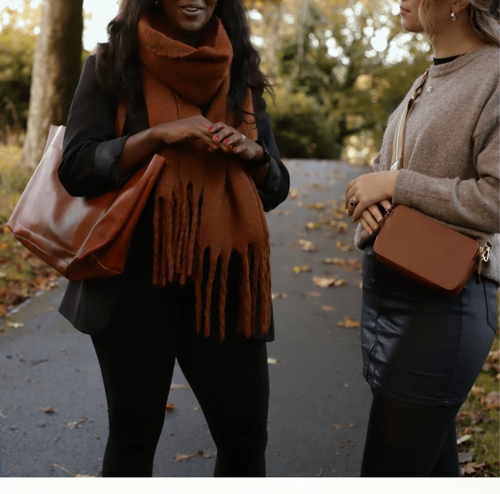 Two women in a park, standing side by side with their bags, amidst a backdrop of trees and grass.