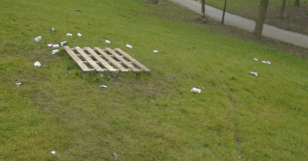 A weathered wooden pallet placed on a grassy area, surrounded by A weathered wooden pallet placed on a grassy area, surrounded by disguarded beer cans.