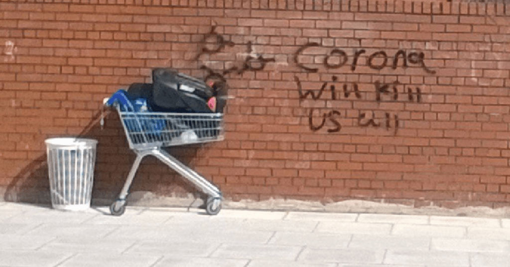 A shopping trolley, abandoned, with waste items when moving house. Graffiti on wall reads "Corona will kills us all".