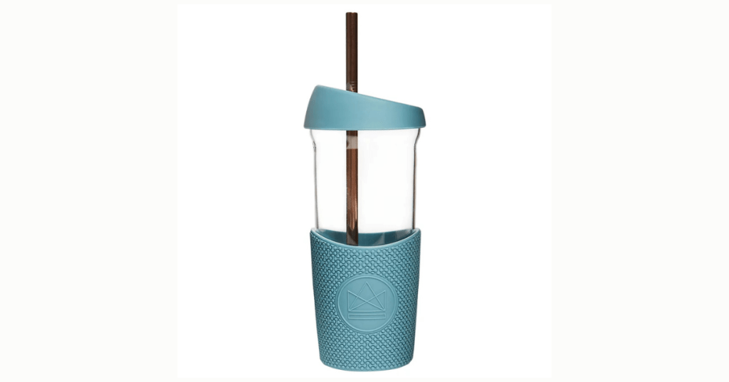 A reusable drinking cup with a transparent body and a blue lid featuring an opening for a straw. A brown straw is inserted through the lid. The lower half of the cup is wrapped in a textured blue grip sleeve that displays a circular emblem with a geometric design. The cup has a sleek, modern appearance and is designed for Eco-friendly use.