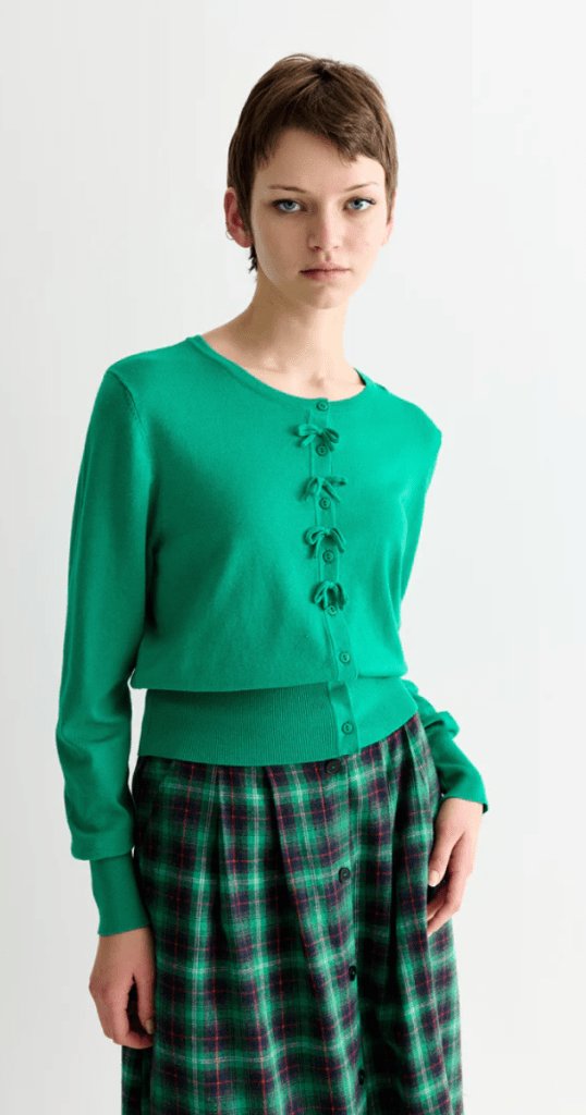 A woman in a green cardigan and plaid skirt smiles, presenting a chic and coordinated look.