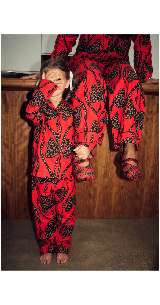 Matching red bow family pyjamas set featuring bold abstract patterns; child barefoot and playfully covering one eye, adult seated on wooden counter wearing coordinating slippers—cosy, stylish loungewear in a warm home setting.
