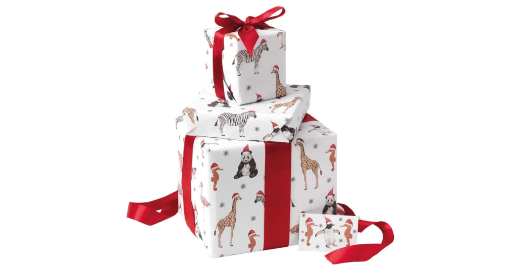Stack of three festive gift boxes wrapped in Christmas animal print recycled wrapping paper featuring zebra, giraffe, panda, flamingo, tiger, and penguin in Santa hats. Each box is tied with bright red ribbon bows. Includes matching gift tag. Cheerful holiday packaging ideal for Christmas presents.
