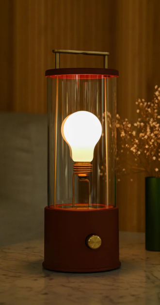 The Muse Portable Lamp with a transparent glass cylinder, glowing bulb-style light core, and deep red base with metallic knob. Features a sleek handle for easy portability and ambient lighting. Stylish and functional, ideal for modern interiors and cozy spaces.