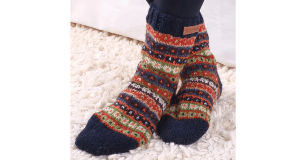 Pair of thick Colorado Sofa Socks with intricate multicolor knit pattern in navy, red, green, orange, and white geometric bands. Featuring solid navy toe and cuff, plus a small brown tag detail. Shown worn on feet resting on a soft white shaggy carpet—ideal for cozy lounging and winter warmth.