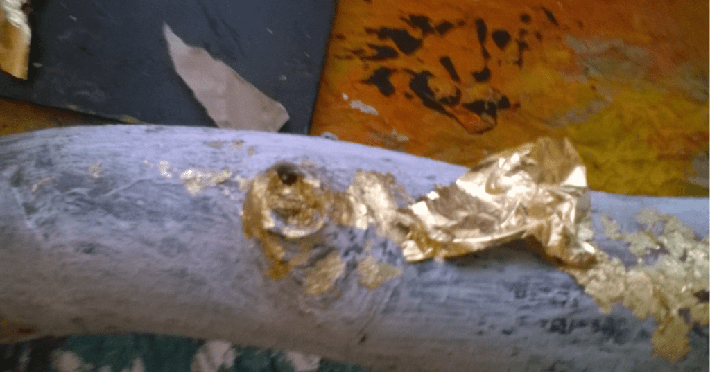 Close-up of a painted surface in pale blue-gray with irregular patches of gold leaf, some loosely adhered, suggesting a work-in-progress gilding technique. In the background, a vivid abstract image of a tiger’s face in orange, black, and yellow adds contrast and energy to the composition.