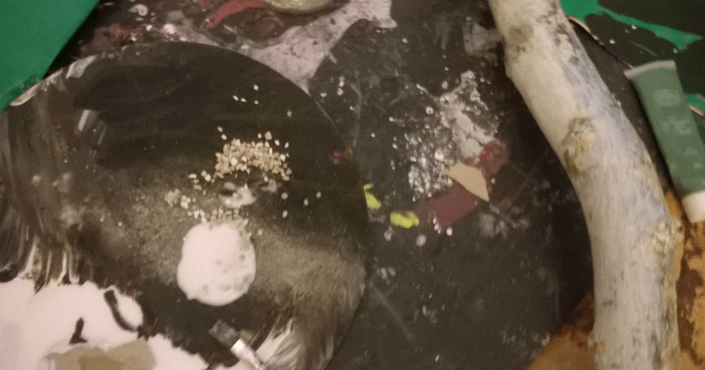 Artist’s workspace featuring a black circular surface with PVA and scattered granular material, sand and pebbles. A paintbrush rests nearby, surrounded by a splattered background of red, yellow, and white paint. A curved, textured wooden piece and a tube of paint, a mixed-media project.