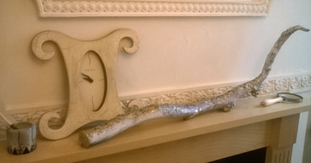 Decorative mantelpiece with a vintage-style oval clock. A painted mug holding a brush sits beside a long, curved decorative tree branch. On the right, a white-handled magnifying glass rests near ornate wall molding, creating a visually rich mix of natural textures and vintage decor.