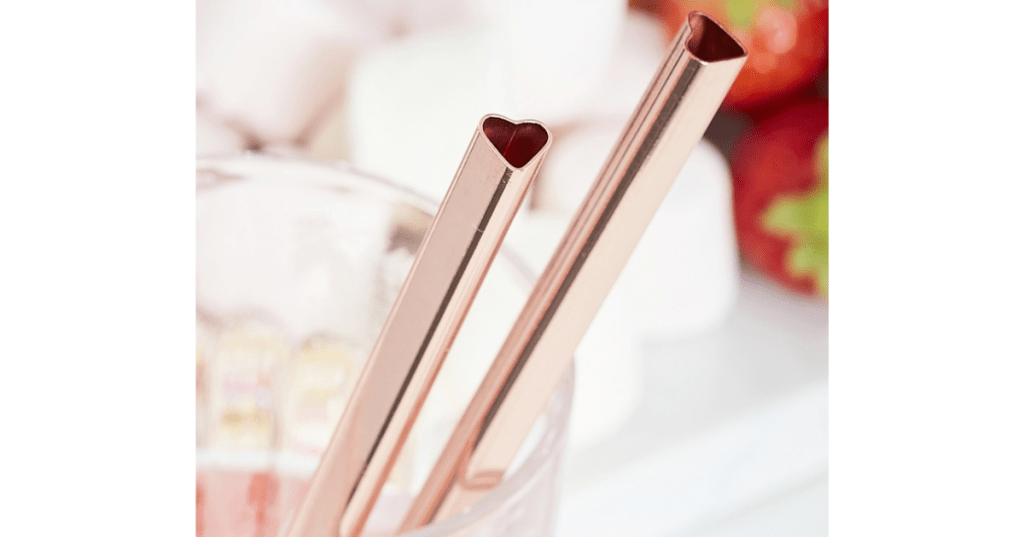 Two rose gold metal straws with heart-shaped openings rest inside a clear glass, set against a soft-focus background of strawberries and marshmallows. The romantic design and warm metallic finish make these reusable straws ideal for Valentine’s Day, weddings, or festive gifting.