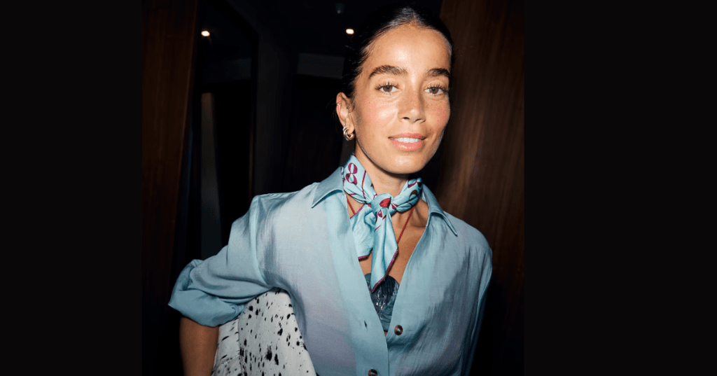 Person wearing a light blue shirt and gold hoop earrings, styled with a bingo-themed bandana featuring a pink number 8. Dimly lit background contrasts with vibrant bandana colours.