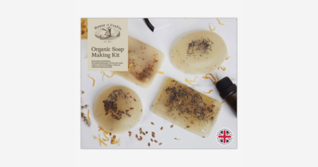 Product packaging for “Organic Soap Making Kit” by House of Crafts. The box displays four handmade soap bars with embedded organic ingredients like seeds and flower petals. A small brown bottle and scattered botanicals surround the soaps. A circular emblem with the British flag indicates the kit is made in the United Kingdom. Multilingual text describes the kit in English, French, German, Spanish, and Portuguese.