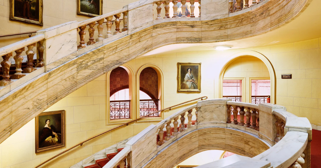 Grand interior of the historic Royal Horseguards Hotel featuring a sweeping marble staircase with ornate balustrades. The staircase curves elegantly across multiple levels. Framed portraits in period attire line the warm-toned walls. Arched windows and doorways with decorative ironwork allow natural light to illuminate the space, highlighting the classical architectural details.