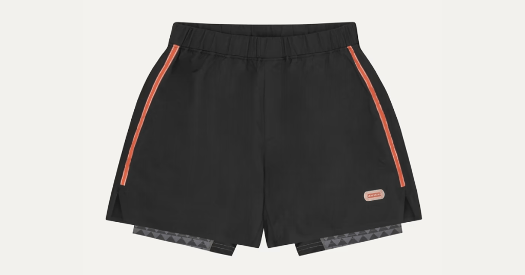 Designer black athletic shorts with orange side stripes and elastic waistband. Features a "Neatishead" logo patch and layered inner shorts with geometric black and grey pattern—premium sportswear with bold, fashion-forward detailing.