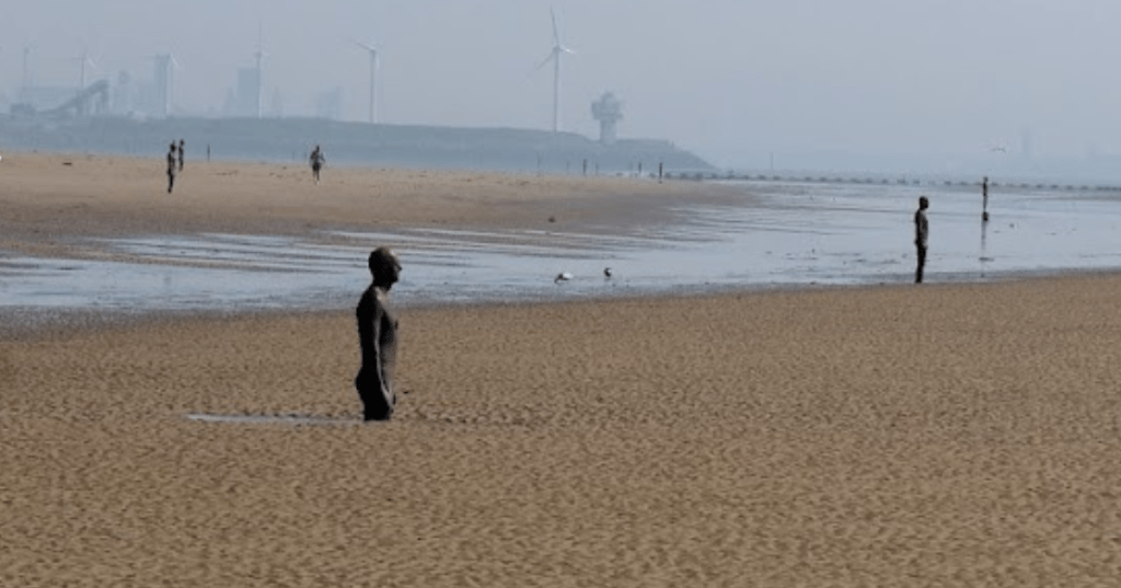 A wide view of Crosby beach in Liverpool with multiple life-sized iron human sculptures standing upright in the sand and shallow water, spaced evenly along the shoreline. Each figure faces out to sea, evoking a sense of solitude and contemplation. In the distance, wind turbines and industrial structures rise against a hazy sky, contrasting the natural and man-made elements. The beach is textured with rippled sand and scattered birds, creating a quiet, surreal atmosphere.