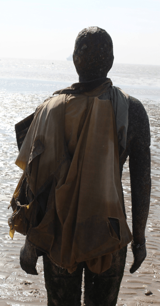 A weathered iron human figure sculpture, created by Antony Gormley, stands on Crosby beach, facing the sea. Its surface is mottled with rust and marine growth, and a tattered piece of fabric is draped over its shoulders, fluttering slightly in the breeze. The figure’s stance is still and solemn, evoking endurance and solitude. Behind it, sunlight glints off the calm water, and the wet sand reflects the soft light, creating a haunting, contemplative atmosphere.