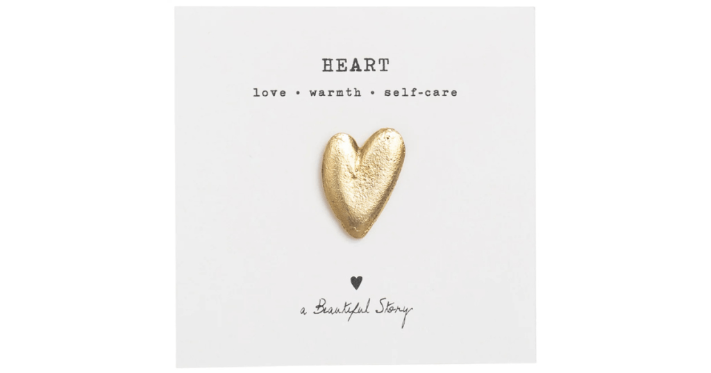 Minimalist card featuring a gold heart-shaped brooch with the word 'HEART' above and the phrase 'love • warmth • self-care' below. Includes a small black heart icon and handwritten text 'a Beautiful Story'. A symbolic gift item promoting emotional well-being and self-care.