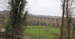 Scenic countryside view from the Llangollen Canal, featuring a grand stone viaduct with multiple arches spanning a green valley. Surrounded by grassy fields, leafless trees, and rolling hills, the tranquil landscape includes a dirt path and bench in the foreground, highlighting the canal’s elevated vantage point.