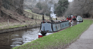 Three narrowboats moored along the Llangollen Canal, including the traditional "Willow Wren Stockton" with chimney smoke rising. A grassy towpath runs alongside the water, opposite a wooded embankment with rustic fencing. The tranquil scene captures canal life in a rural setting with hints of winter.