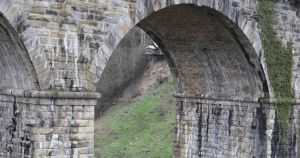 Close-up view of the Chirk Viaduct, showing two large stone arches built from weathered rectangular blocks. Ivy climbs part of the structure, and a grassy slope with a small wooden shelter is visible through the central arch. The scene blends historic engineering with natural surroundings.