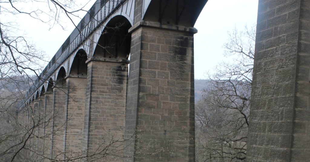 Pontcysyllte Viaduct spanning a wooded valley, with tall stone piers supporting a metal aqueduct channel. The structure’s arches rise above leafless trees and rolling hills, showcasing historic engineering in a winter landscape.