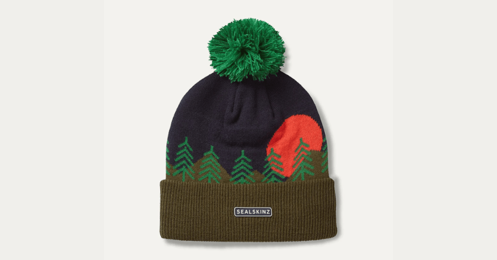 Knitted winter beanie hat with pom-pom, featuring a nature-themed design of green pine trees and a large red sun or moon on a dark blue background. Ribbed olive green brim with 'SEALSKINZ' label. Stylish and warm outdoor accessory for cold weather and nature lovers.
