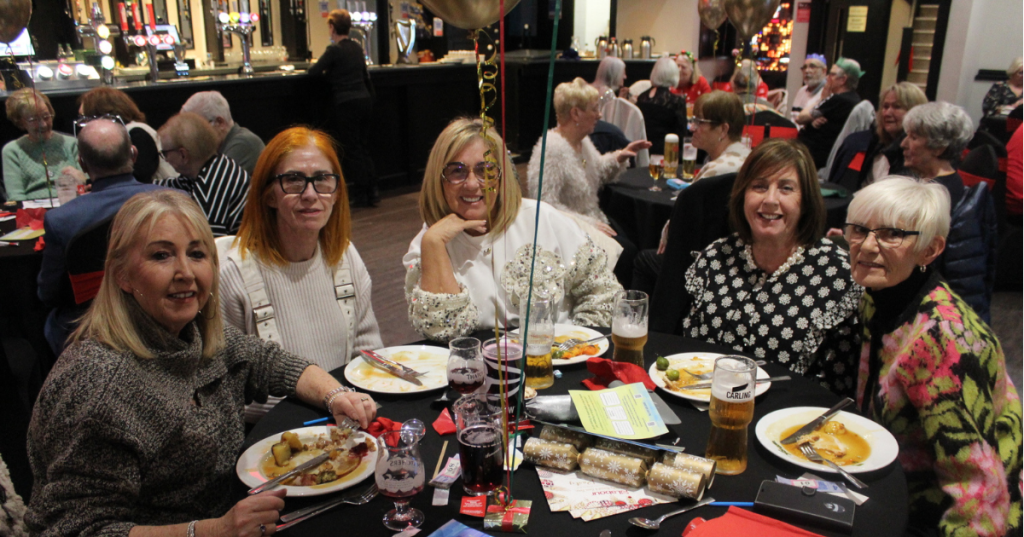 A lively holiday celebration at a the Sandon, with guests seated at round tables enjoying a festive meal. In the foreground, six women sit together surrounded by plates of food, drinks, and Christmas-themed decorations including balloons and crackers. The background shows more attendees chatting and dining, creating a warm, communal atmosphere.