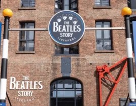 Arriving at The Beatles Story museum in Liverpool, visitors are greeted by a historic red brick building with arched windows and a bold circular sign featuring the faces of John, Paul, George, and Ringo. Below the sign, “THE BEATLES STORY LIVERPOOL” is displayed in crisp white lettering on the wall. The entrance evokes the city’s dockside heritage, with a red metal structure to the right hinting at its maritime past. The museum promises an immersive journey into the legacy of The Beatles, set against the backdrop of Liverpool’s iconic waterfront.