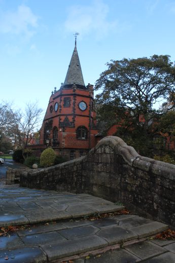 Historic brick building with clock tower viewed from behind a stone bridge in Port Sunlight. Autumn leaves scatter the walkway, and sparse trees frame the scene, highlighting the village’s architectural charm and seasonal atmosphere.