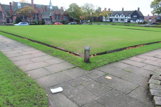 View of the bowling green in Port Sunlight, bordered by low wooden edging and surrounded by paved walkways. Historic brick and timber buildings frame the background, while a small signpost marks the corner of the green, capturing the village’s heritage charm and communal atmosphere.