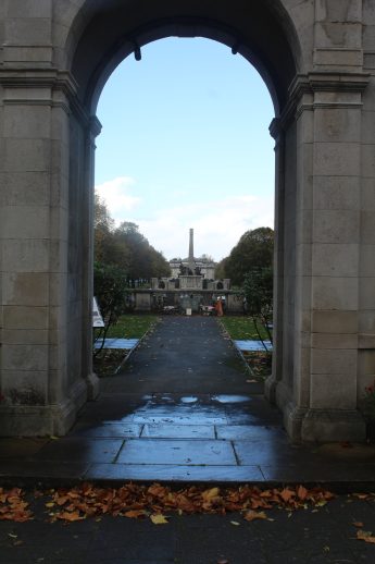 View through a stone archway framing the formal approach to the war memorial in Port Sunlight. A central path leads to a tall obelisk monument surrounded by landscaped gardens and trees. Wet ground and scattered autumn leaves evoke a reflective, seasonal atmosphere.