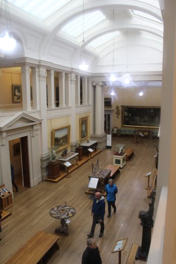 Interior view of the Lady Lever Gallery, showcasing classical architecture with vaulted ceilings, columns, and ornate moldings. Artworks including paintings and sculptures are displayed throughout the well-lit space, as visitors explore the exhibits in this historic cultural setting.