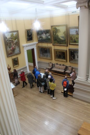 Group of visitors, including children, gathered inside the Lady Lever Gallery beneath high ceilings and classical white columns. Framed paintings line the walls, and antique furniture accents the wooden floor, creating an elegant setting for art appreciation and cultural learning.