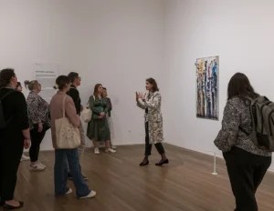 Visitors gather around a Tate Modern guide during the Official Discovery Tour, engaging with a bold abstract painting from the "Painterly Gestures" exhibition. The guide gestures expressively, sharing insights into the artwork’s technique and meaning. Set in a minimalist gallery with wooden floors and white walls, the scene captures cultural immersion and educational value.