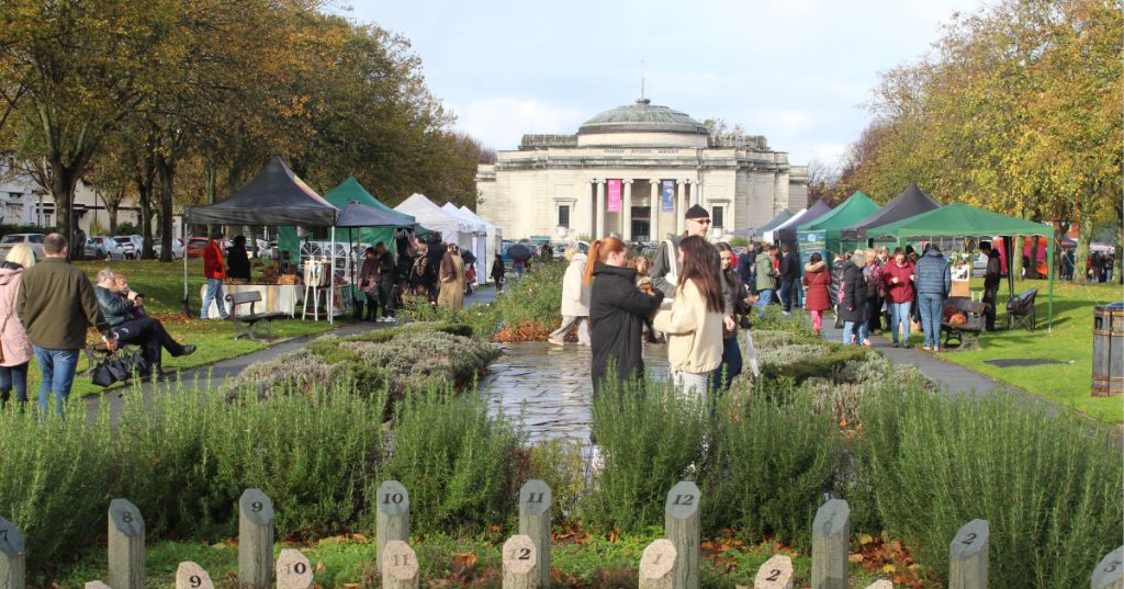 A crowd of people navigates a park filled with greenery, close to the Lady Lever Gallery during a lively art market event.