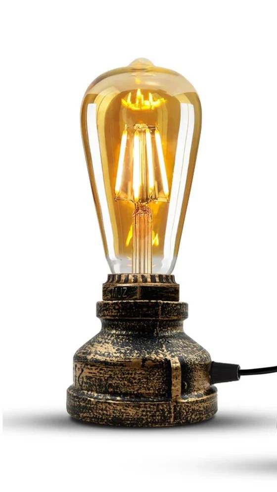 Vintage-style industrial table lamp with exposed filament bulb emitting warm yellow light. Features a distressed metal base, inline switch on black cord, and UK three-pin plug. Ideal for ambient lighting in retro, rustic, or eclectic interiors.