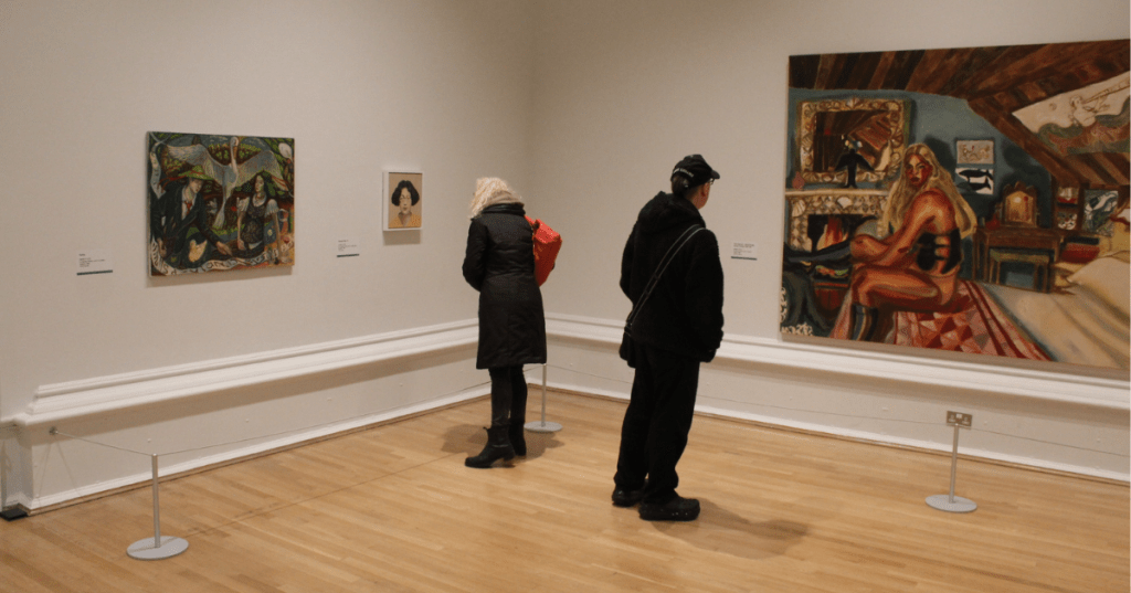 Two visitors explore the John Moores Painting Prize 2025 exhibition in a minimalist gallery space. One person, wearing a red backpack, closely examines a small portrait, while another observes a large figurative painting of a woman seated in a domestic interior. The white walls and wooden floor create a calm setting for art appreciation.