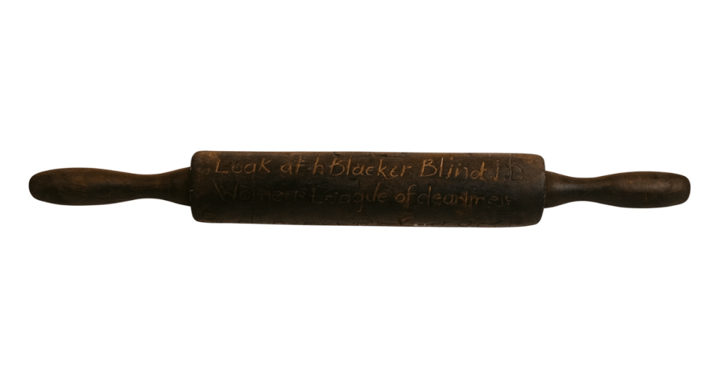 Art piece, hand engraved rolling pin made of dark, weathered wood, isolated against a white background. Features hand-carved inscriptions including “clean my cupboards once a week,” “Look at how Black her Blind is,” and “Women’s League Task of cleanliness.” The rolling pin has tapered handles and visible wear, suggesting historical or symbolic significance related to domestic labour and social commentary.