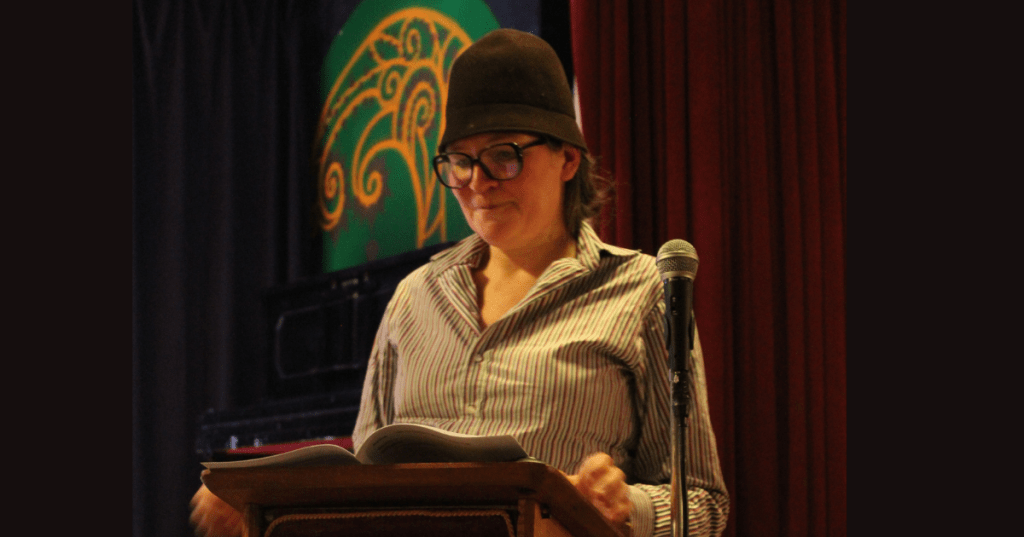 Alison Little, dressed as a Suffragist, reading Between the Pages—her own work—at the People of Anfield celebration held at the Liverpool Irish Centre. She stands at a podium with microphone, wearing a striped shirt, glasses, and brown hat, framed by a red curtain and swirling green-and-yellow backdrop. A moment of heritage storytelling and community performance.
