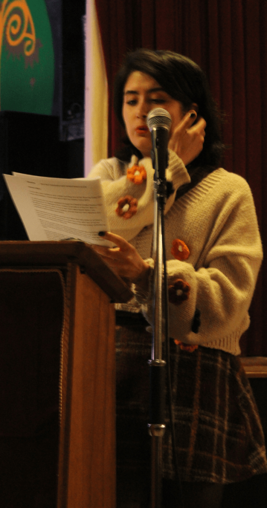 Farida Bamyanchi reading at the People of Anfield celebration, Liverpool Irish Centre. She stands at a wooden podium with microphone, holding a printed document, wearing a beige floral sweater and plaid skirt. Set against a red curtain and decorative green backdrop, the image captures a moment of spoken word performance and community storytelling.