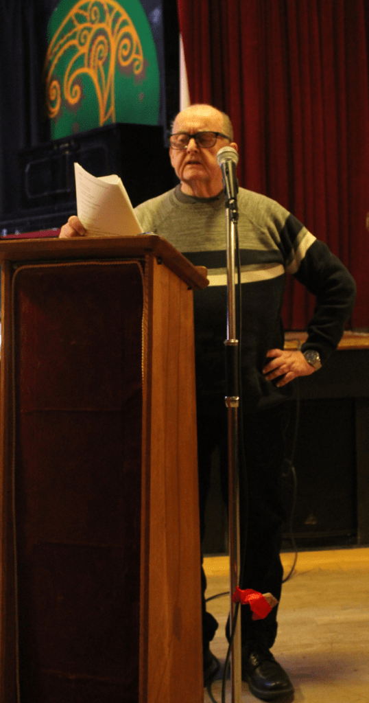 Ian Watt reading at the People of Anfield celebration, Liverpool Irish Centre. He stands at a wooden podium with microphone, holding a sheet of paper, dressed in a dark sweater with a white stripe. The stage is framed by red curtains and a green backdrop featuring a stylized tree design, capturing a moment of community storytelling and local heritage.
