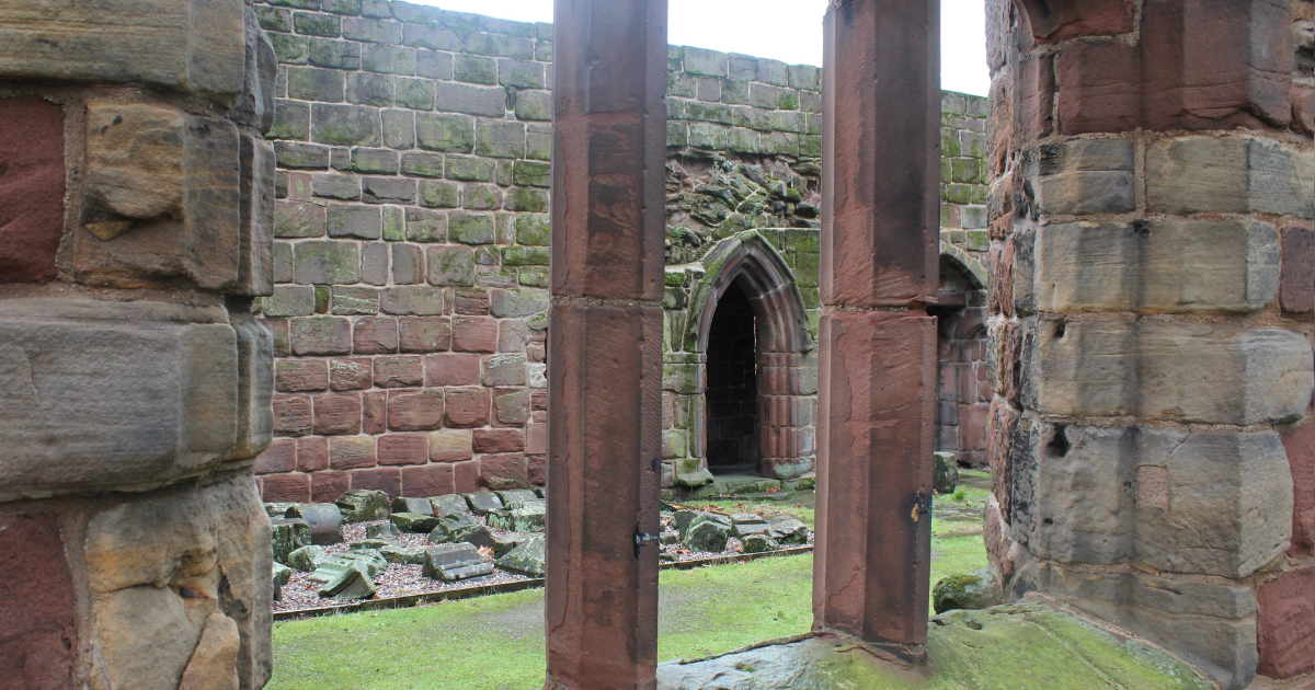 View through three vertical stone columns framing the interior ruins of Birkenhead Priory. The moss-covered courtyard and scattered stone debris lead to a reddish-brown stone wall with a pointed Gothic arch doorway, showcasing medieval craftsmanship and architectural decay. A quiet glimpse into England’s oldest surviving Benedictine monastery.