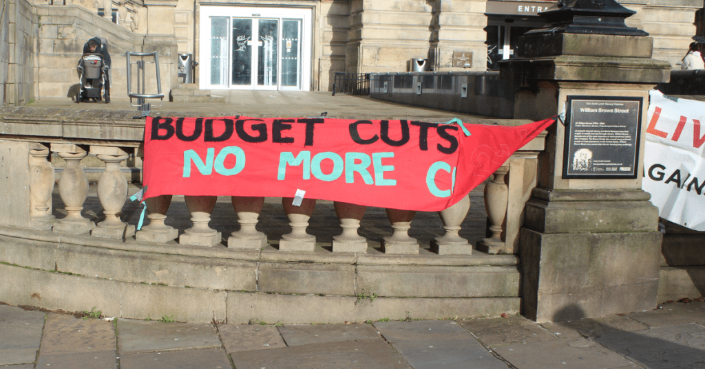 Red protest banner reading “BUDGET CUTS NO MORE C…” draped over a stone balustrade on William Brown Street in Liverpool, during a demonstration against the closure of Breck Road Library. The historic setting and visible plaque emphasize the cultural significance of libraries and public funding.