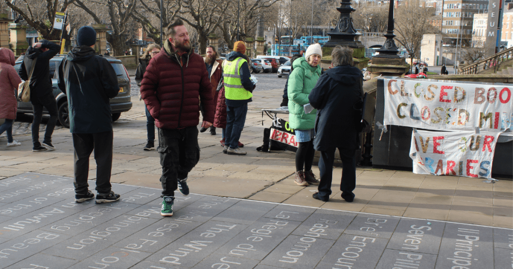Public protest in Liverpool against the closure of Breck Road Library, with demonstrators gathered near the World Museum entrance. A handmade banner with partially obscured text—“CLOSED BOOKS CLOSED MINDS SAVE OUR LIBRARIES”—is displayed, emphasizing community resistance and the cultural importance of public libraries. Engraved literary quotes on the pavement add symbolic weight to the setting.
