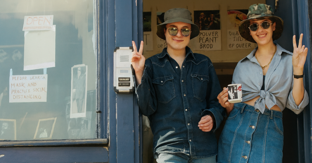 Tracey Emin and Sarah Lucas stand in the doorway of The Shop, flashing peace signs in casual denim outfits and sunglasses. Their relaxed, rebellious energy reflects the spirit of their 1990s East London art collaboration. Behind them, posters opposing the death penalty and an “OPEN” sign hint at The Shop’s role as both creative space and activist hub. A candid moment capturing feminist defiance, DIY aesthetics, and the raw charisma of two Young British Artists reshaping the art world.