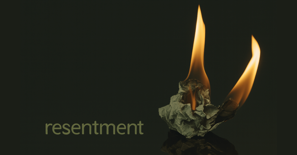 A crumpled piece of paper labeled “resentment” burns against a solid black background. Two vivid flames rise from the paper’s edges, glowing orange and yellow, casting warm light and subtle reflections on the dark surface. The word “resentment” appears in lowercase greenish-gray text on the left side of the image, slightly textured and evenly spaced. The composition is stark and symbolic, evoking emotional release and transformation through fire.