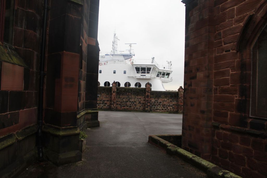 View from Birkenhead Priory toward the Cammell Laird docks, where a large white ferry is partially visible between two dark stone buildings. The ship contrasts with the historic architecture, highlighting the layered industrial and monastic heritage of the Mersey waterfront.