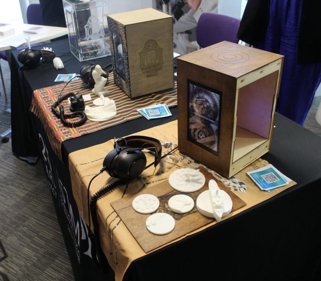 Interactive “time machine” display created by Liverpool School of Tropical Medicine; features wooden touchscreen boxes, 3D-printed models including ships and seated figures, headphones for audio storytelling, and QR codes for digital content—designed to explore history, science, and global health through tactile and multimedia engagement.