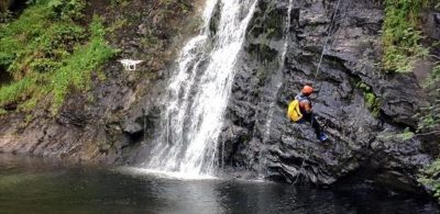A climber wearing a helmet and harness abseils down a rocky cliff beside a waterfall, with a drone hovering nearby, capturing the action. The lush greenery and rugged terrain evoke the dramatic landscapes associated with adventure sports in Snowdonia.