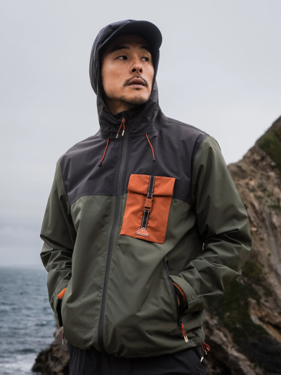 Person standing on a rocky coastline wearing a Burlingham Waterproof Packable Jacket, showcasing its hooded design, colour‑blocked panels, and weather‑resistant features in an outdoor setting.